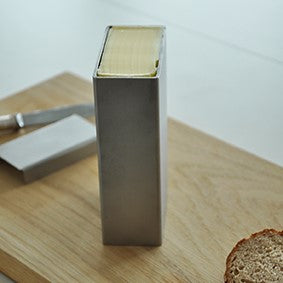 Butterdose aus Edelstahl mit rechteckigem Design, gefüllt mit Butter, auf einem Holzbrett neben einer Brotscheibe